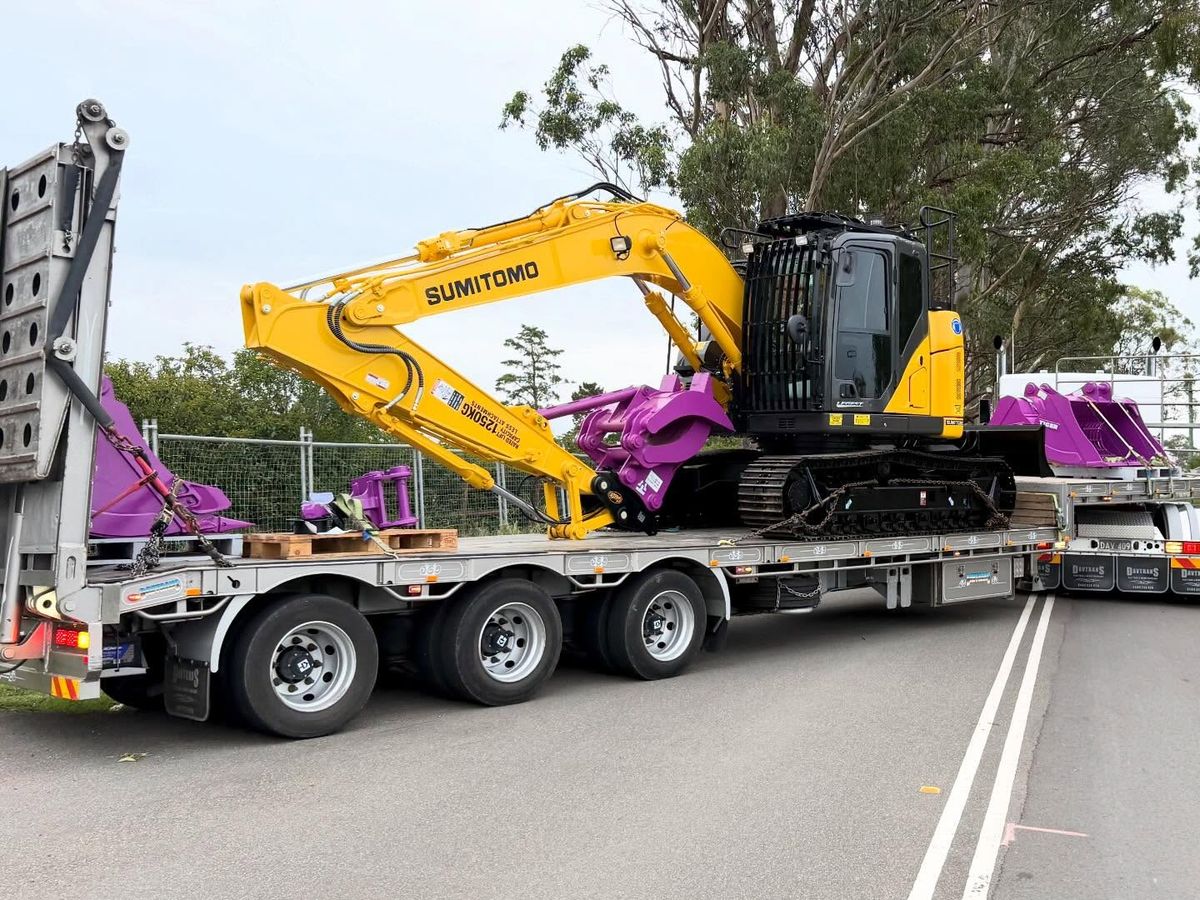 Yellow Sumitomo excavator with purple attachments on a float trailer