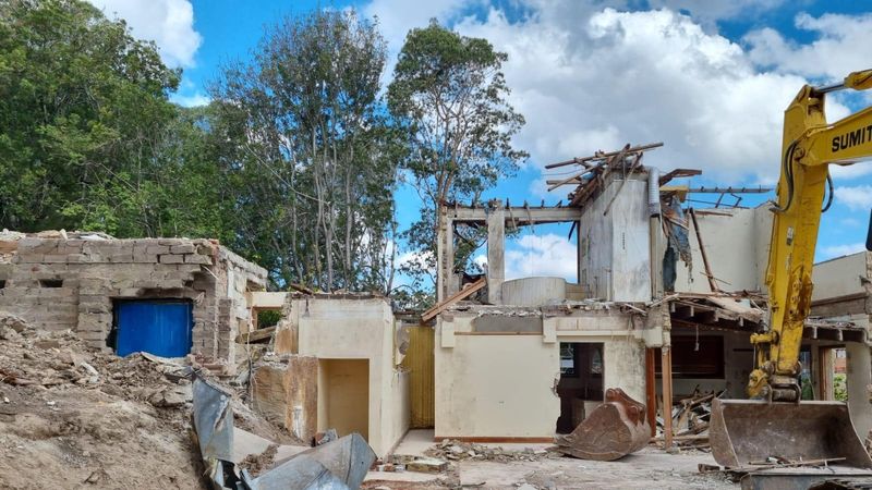 House demolition in progress: excavator breaking through remaining walls with roof already removed