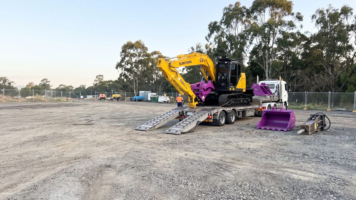 Excavator hire in Sydney: yellow excavator with purple bucket and hammer attachments on a float trailer