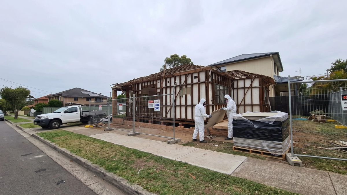 Asbestos removal in Sydney: workers in white protective suits at a fenced house site with wrapped sheeting stacked ready